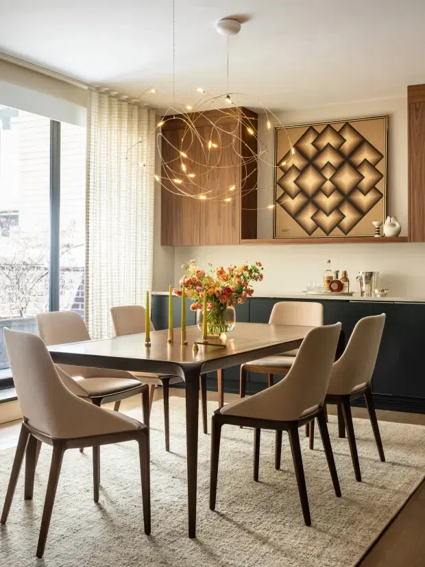 A refined dining room with a rectangular dark wood dining table surrounded by upholstered dining chairs, set on a large neutral rug. Floor-to-ceiling glazing with sheer curtains brings in abundant daylight. The room includes dark built-in cabinetry, a warm wood wall panel, geometric framed artwork, a sculptural cluster pendant light, and a floral centrepiece.