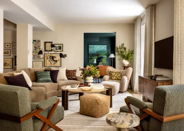 Warm, refined living room with a large neutral sectional sofa, mid-century-inspired lounge chairs, a pair of wood-framed armchairs, round coffee tables, and a leather ottoman. The room features cream walls, full-height textured curtains, a dark timber media console, curated artwork, indoor plants, and a blue-toned alcove or view beyond the room. The overall composition is layered, comfortable, and sophisticated.