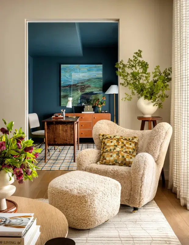 A warmly styled living room with a plush cream boucle lounge chair and matching ottoman in the foreground, set on a pale patterned rug. A round wood coffee table, leafy plants, textured curtains, and a patterned accent cushion add softness and colour. Through a wide opening is a dark blue home office or study with a wooden desk, cabinetry, chair, desk lamp, and artwork. The space combines warm natural timber, deep blue walls, soft upholstery, and eclectic mid-century-inspired furnishings.