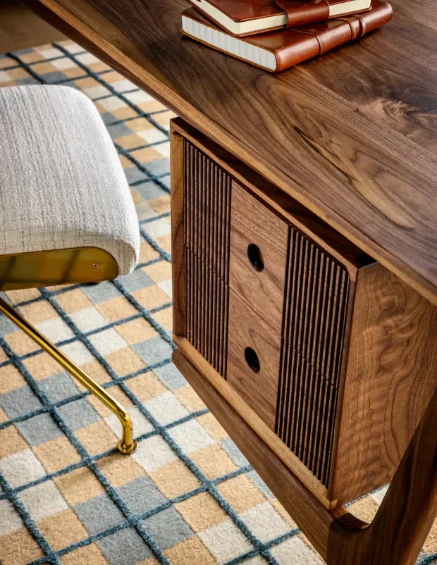 Close-up interior detail of a mid-century-style timber sideboard or credenza with richly grained walnut cabinetry, vertically fluted doors, and dark circular pulls. A cream upholstered chair with a brass-coloured metal leg sits beside it on a geometric cream, grey, and blue grid-pattern rug. Leather-bound books rest on the furniture surface.