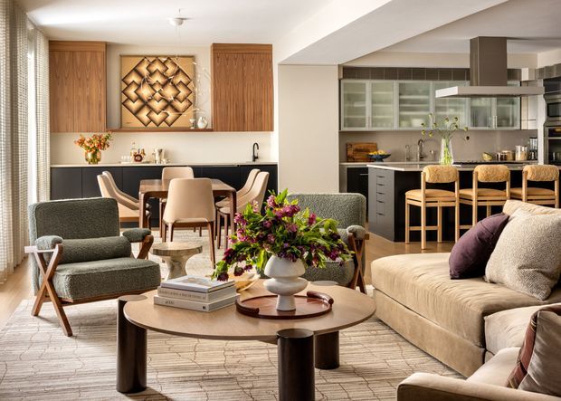 Open-plan contemporary living, dining, and kitchen interior with warm timber cabinetry, a dark kitchen island, upholstered seating, and a layered neutral palette. The living area features a large sectional sofa, accent chairs, nested round coffee tables, a patterned rug, and a floral arrangement. A dining table and chairs sit behind the lounge, with the kitchen visible beyond.