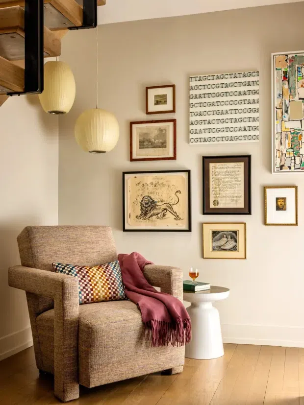 A warm, curated living room corner featuring a textured upholstered armchair, patterned cushion, burgundy throw, small round side table, pendant lights, timber flooring, and a gallery wall of framed artwork and prints. A timber stair or shelving structure is visible at the upper left.