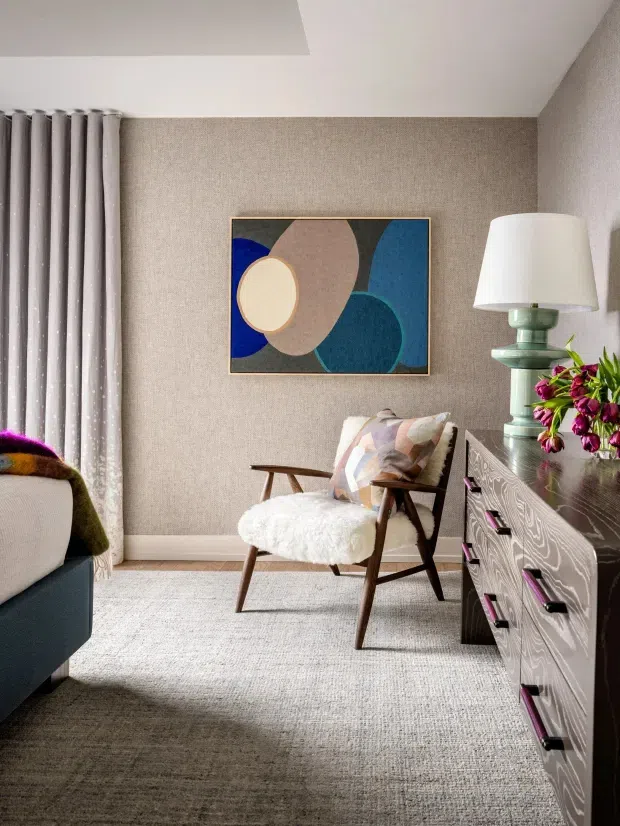 A warmly styled sitting area featuring a mid-century-inspired upholstered armchair, textured neutral wall covering, abstract geometric artwork, a sculptural table lamp, patterned dresser, soft curtains, and a pale textured rug. The edge of a sofa is visible at left.