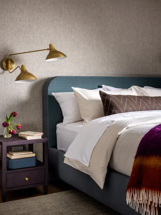 A warmly styled bedroom featuring a deep teal upholstered bed with white layered bedding, a patterned brown accent cushion, and a dark bedside table with books and flowers. Brass wall-mounted lamps illuminate textured grey-beige wallcovering, while a rich burgundy and purple throw adds colour in the foreground.