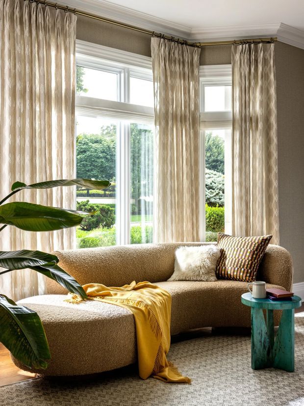 A bright, refined living room with a curved textured beige sectional sofa positioned in front of tall bay windows. Floor-to-ceiling patterned curtains, white trim, a neutral patterned carpet, tropical foliage, a mustard throw, and a small turquoise side table create a warm contemporary setting with abundant natural light.