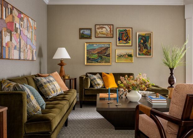 A richly layered living room with olive green velvet sofas, patterned cushions, a dark wood coffee table, and a pair of upholstered armchairs. The taupe walls are arranged with a gallery of framed artwork, complemented by a large abstract wall textile, table lamps, and floral styling. Traditional detailing, warm timber tones, and an eclectic mix of colour create a refined yet comfortable atmosphere.
