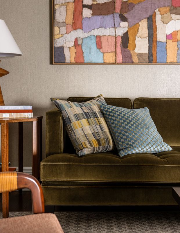 Close-up interior view of a refined living room featuring a deep olive-green velvet sofa, patterned blue and multicoloured cushions, a textured neutral wall, framed abstract artwork, a wooden side table with books, and a floor lamp. The space has a warm, richly layered and eclectic mid-century-influenced character.