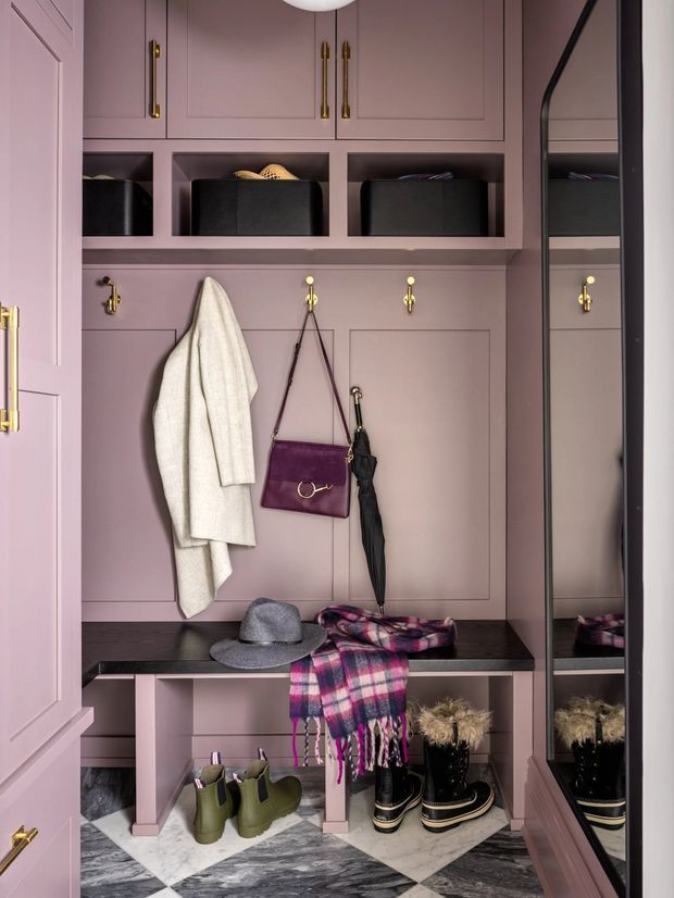 A compact, elegantly fitted entryway or mudroom with dusty mauve built-in cabinetry, upper storage cubbies, paneled doors, brass hooks and hardware, a dark bench seat, and a full-height mirror. Coats, handbags, umbrellas, hats, scarves, and boots are organised within the joinery. The floor features a bold grey-and-white geometric marble-like checkerboard pattern.