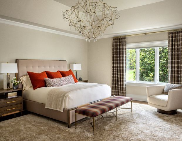 Elegant bedroom with a tufted upholstered bed, layered white and rust-red bedding, dark bedside cabinetry, a patterned upholstered bench, a cream armchair, full-length plaid curtains, and a large window overlooking greenery. The room has a soft neutral palette, plush carpeting, decorative ceiling molding, and a sculptural contemporary chandelier.