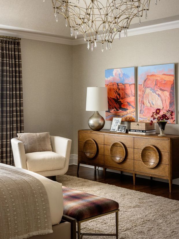 Refined sitting room with a warm neutral palette, textured beige walls and carpet, dark timber flooring, full-height patterned curtains, and a sculptural brass-and-crystal chandelier. A cream upholstered armchair and sofa surround a patterned ottoman, while a mid-century-inspired timber sideboard with circular carved doors displays colourful abstract artwork and decorative objects.