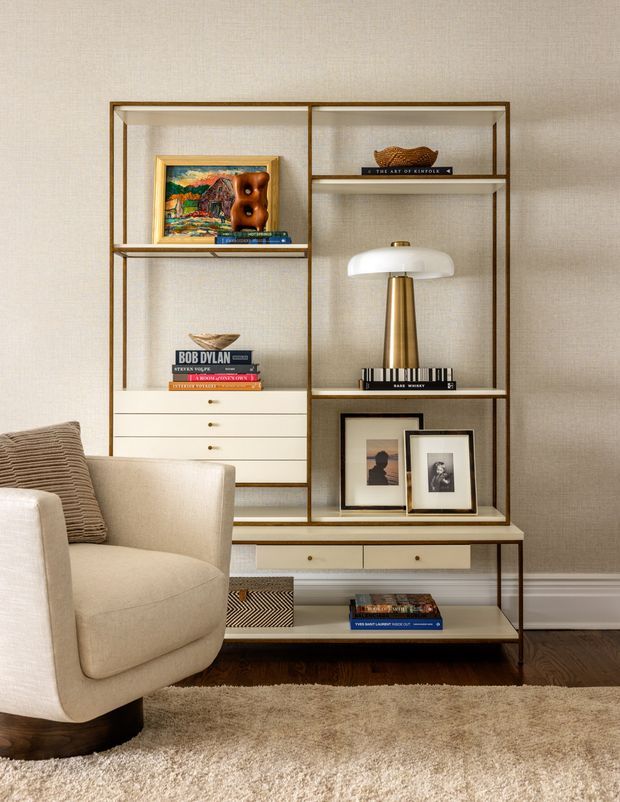 A refined living room vignette featuring a large open timber-and-metal shelving unit with integrated drawers and a console surface. The shelves display books, framed artwork, ceramics, and a sculptural brass-and-white table lamp. A cream upholstered armchair sits in the foreground on a deep-pile neutral rug, against textured beige wallcovering with white trim and dark timber flooring.