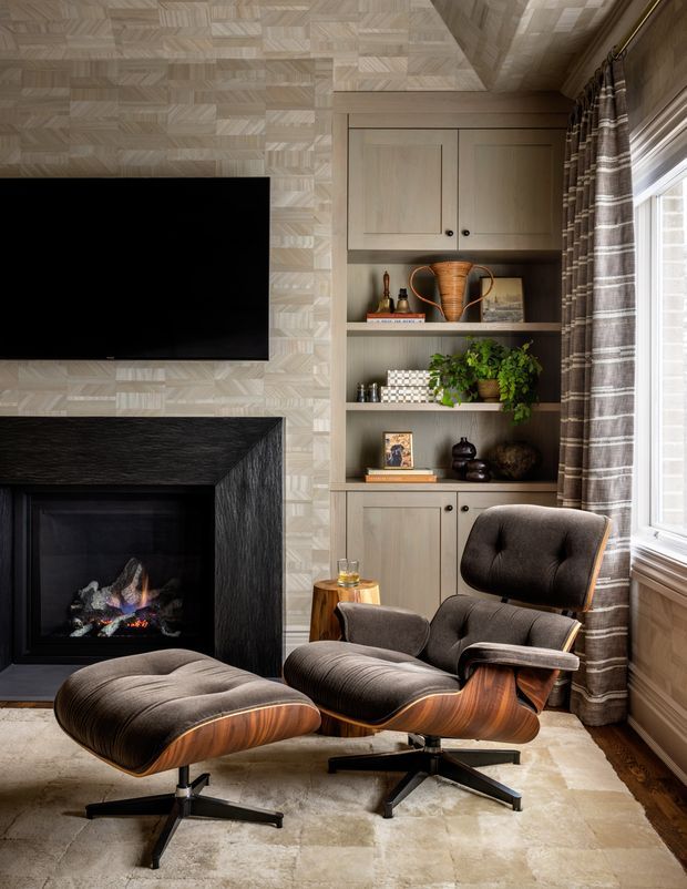 A refined living room with a wall-mounted television above a dark textured fireplace, integrated light-wood shelving and cabinetry, and a mid-century-style lounge chair with matching ottoman. Patterned wall covering, layered curtains, a neutral rug, and warm wood details create a cosy, sophisticated interior.