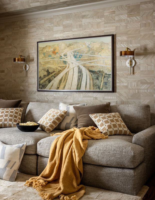 A warm, richly layered living room centered on a large textured grey sectional sofa. The room features a geometric light timber wall treatment, a framed landscape artwork, symmetrical brass wall sconces, patterned cushions, and a mustard throw draped over the sofa. The overall composition is cozy and refined with strong geometric patterning and natural neutral tones.