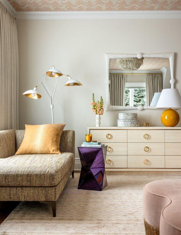 A refined living room with a textured upholstered chaise sofa, warm gold cushion, sculptural multi-arm floor lamp, pale wood dresser, oversized decorative mirror, and rounded upholstered ottoman. The room features soft neutral walls, patterned wallpaper on the ceiling, full-height curtains, layered carpet, and warm brass and amber accents.