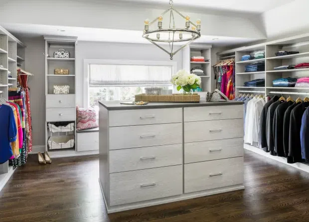 Bright, spacious walk-in wardrobe with built-in open shelving and hanging rails on three sides, a large central light-wood island dresser, dark timber flooring, a window with a sheer blind, and a decorative chandelier. Clothing, shoes, and accessories are neatly organised throughout the fitted storage.