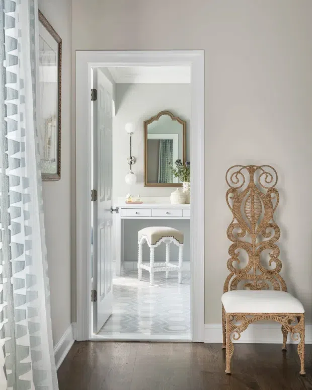 Bright, refined interior view through an open doorway into a light-filled dressing or vanity area. The foreground has dark timber flooring, pale walls, patterned sheer curtains, a woven ornate accent chair, and framed artwork. Beyond the doorway is a white built-in vanity with drawers, a small upholstered stool, decorative mirrors, pendant-style wall lighting, and patterned pale flooring.