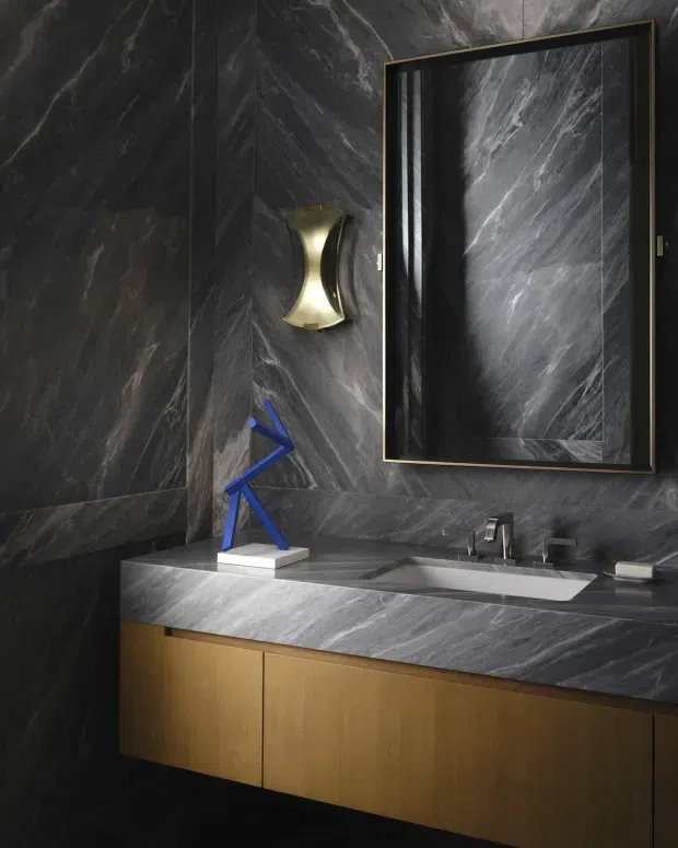 A dramatic, luxurious bathroom vanity with dark veined stone wall cladding and matching stone benchtop, a wide integrated white basin, brushed metal tapware, and warm timber floating cabinetry. A large black-framed mirror with a slim brass-toned edge dominates the wall, complemented by a sculptural brass wall light and blue decorative object. The space has a moody, high-contrast contemporary look.