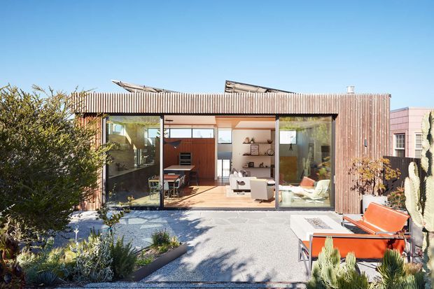 Single-storey modern dwelling viewed from a landscaped outdoor area, with a flat-roofed timber-clad facade and extensive floor-to-ceiling glazing opening onto a paved courtyard or patio. The interior is visible through the glazing, including a living area and kitchen/dining zone. The exterior uses warm vertical timber cladding and a restrained contemporary composition, while bright orange outdoor seating provides a small accent.