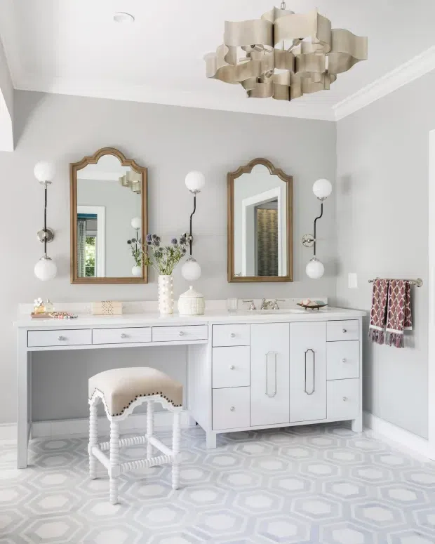 Bright, refined bathroom vanity area with a long white double vanity, integrated countertop basins, decorative framed mirrors, globe wall sconces, patterned geometric floor tiles, and a sculptural multi-panel ceiling light. The room uses pale grey walls, white cabinetry and trim, and subtle brass-toned accents.