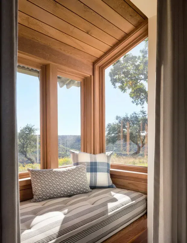 A compact built-in window seat occupies a bright corner framed by extensive warm timber panelling, trim, and exposed ceiling boards. Large timber-framed windows overlook a wooded, hilly landscape. The seat has a striped upholstered cushion and patterned and blue-check pillows, creating a cosy reading nook with abundant natural light.