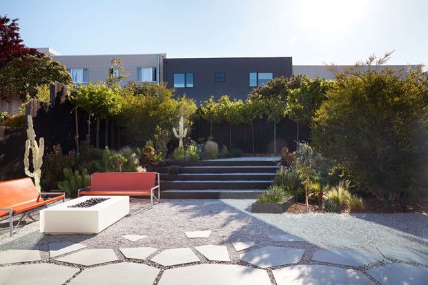 A landscaped contemporary outdoor courtyard or terrace with a gravel-paved surface, irregular large stepping stones, raised planting beds, mature shrubs and succulents, and broad steps leading toward a modern dark-clad house. Bright outdoor lounge seating and a rectangular fire pit or feature table create a social entertaining area. The scene has clean geometric architecture softened by lush, drought-tolerant planting.