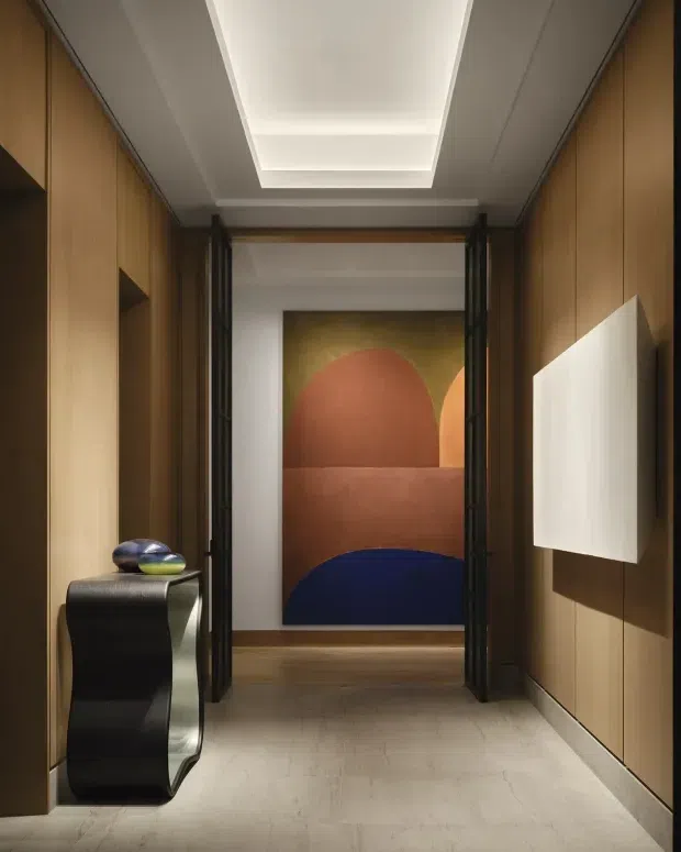 A narrow, refined interior hallway lined with warm timber wall panelling and integrated recessed shelving. A sculptural black console sits along the left wall, while dark-framed glazed or open folding doors lead toward a room displaying a large abstract artwork in terracotta, ochre and deep blue. The corridor has a pale stone-like floor, a white recessed tray ceiling with concealed lighting, and a contemporary, gallery-like atmosphere.
