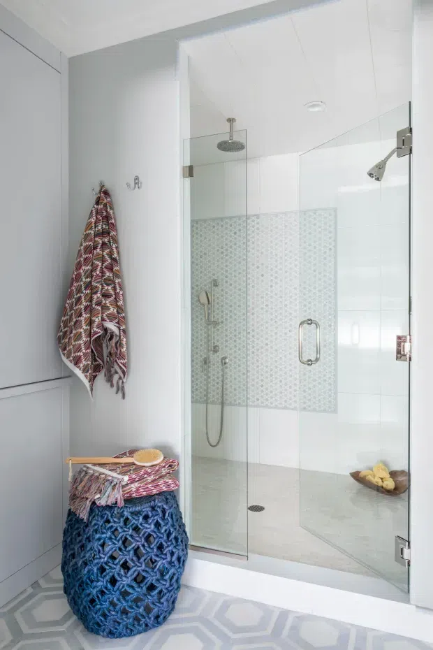 Bright contemporary bathroom featuring a large frameless glass walk-in shower with dual shower fixtures, pale patterned wall tile, white tiled surfaces, and a light geometric floor tile. Built-in white cabinetry occupies the left side, with a textured blue woven stool or basket and patterned textiles adding colour and texture.