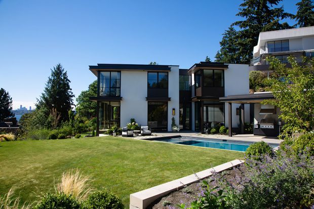 A modern multi-level residential home viewed from a landscaped backyard, featuring crisp white rendered walls, dark-framed floor-to-ceiling glazing, projecting flat rooflines, covered outdoor entertaining areas, and a long rectangular swimming pool beside a broad lawn. Mature planting and ornamental garden beds surround the property, with a distant urban skyline visible beyond the site.