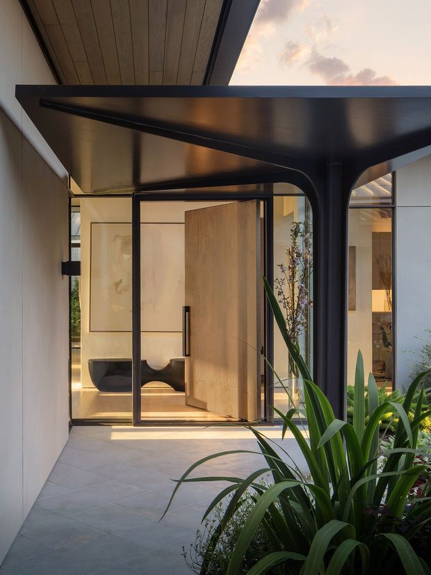 A contemporary covered entry courtyard viewed from outside, with a dramatic dark metal flat canopy supported by sculptural curved columns. Large glass entry doors and a warm timber pivot door open toward a softly lit interior, while lush planting borders the paved approach. The composition contrasts black metal, pale walls, timber ceiling lining, and warm interior light.