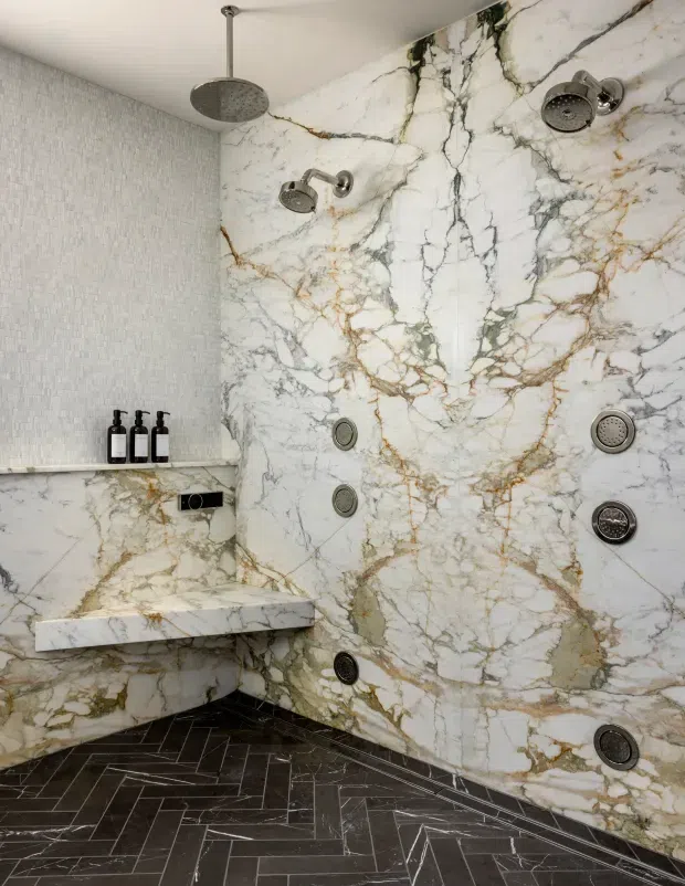 Luxurious walk-in shower with full-height dramatic white marble slab walls veined in grey, black, and warm gold. The shower includes multiple overhead and wall-mounted fixtures, recessed controls, a built-in marble bench, a narrow mosaic-tiled side wall, and a dark herringbone tile floor with contrasting grout.
