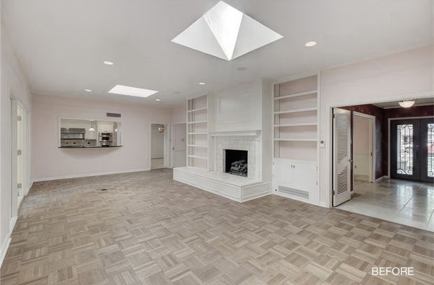 Bright, mostly empty traditional living room viewed toward a white fireplace and built-in shelving. The room has pale walls, recessed ceiling lights, two skylights, and a patterned light-toned parquet or carpet-like floor. A pass-through opening reveals a compact kitchen area on the left, while an open doorway on the right leads to a tiled entry hall with glazed double doors. The image is labelled “BEFORE,” suggesting a renovation context.