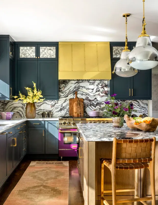 A richly detailed kitchen with deep navy cabinetry, dramatic white-and-grey veined marble splashback and benchtops, and a large island. A polished brass rangehood, brass hardware, and oversized pendant lights add a glamorous focal point, while a timber bar stool, patterned rug, florals, and colourful accessories bring warmth and character.