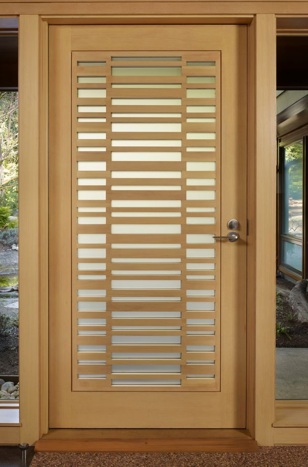 Close-up view of a light timber entry door with a large inset panel of horizontal slatted openings and translucent glazing. The door is framed by matching timber trim and sidelights, with a simple silver lever handle and lock. Exterior landscaping is visible through the glazing, while the interior has a neutral carpeted threshold.