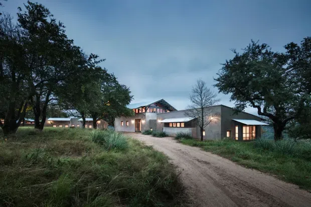Exterior dusk view of a sprawling low-rise contemporary building set within a natural, wooded landscape. The complex has multiple connected volumes with dark pitched roofs, gabled glazed sections, warm interior lighting, and textured grey masonry or concrete walls. A dirt driveway leads toward the entrance, with mature trees and native grasses surrounding the property.