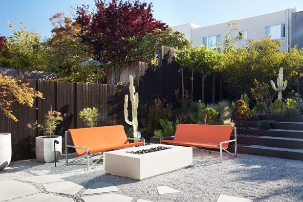 A contemporary landscaped courtyard or outdoor entertaining area with orange outdoor lounge seating arranged around a rectangular fire pit. The space features pale gravel, concrete stepping slabs, dark timber fencing, raised planted garden beds, cacti and sculptural planting, with a modern light-coloured multi-storey building visible beyond.