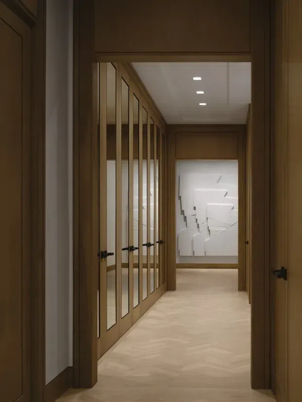A refined residential hallway lined with warm dark-wood cabinetry and multiple glass-panel timber doors. The corridor has a pale herringbone timber floor, recessed ceiling lights, and a white sculptural wall artwork at the far end. The composition emphasizes symmetry, built-in joinery, and a calm, gallery-like transition space.