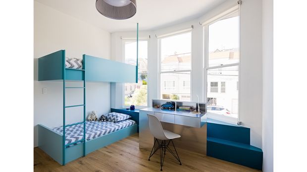 Bright, compact children's bedroom with a built-in teal bunk bed and integrated storage, a matching built-in window seat, and a small study desk positioned beneath tall windows. The room has white walls, warm timber flooring, blue and white patterned bedding, and a modern pendant light. The built-in furniture makes efficient use of the narrow room while allowing abundant natural light.