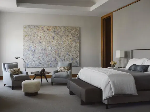 A refined contemporary bedroom with a large upholstered bed, dark grey bed base, white bedding, and a matching bench at the foot. Two upholstered lounge chairs and a small round coffee table form a sitting area beside a textured abstract artwork. The room has neutral grey and white finishes, timber-trimmed recessed door or wardrobe panels, soft carpet flooring, and a recessed tray ceiling with understated lighting.