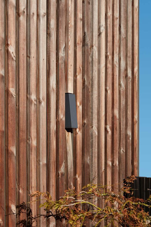 Close-up exterior view of a tall facade clad in vertically arranged weathered timber boards, with a dark rectangular wall-mounted fixture and planting at the base. A clear blue sky is visible along the right edge. The image emphasizes the natural wood texture and vertical cladding pattern.