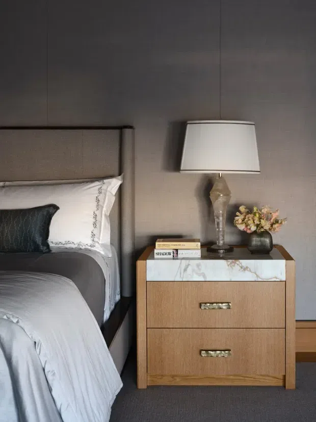 Close-up of a refined bedroom corner featuring a partially visible upholstered bed with grey bedding and a dark accent pillow, a textured grey wall, and a timber bedside table with a white veined stone drawer front. The table holds a sculptural glass lamp, books, and a small floral arrangement. The palette is muted and sophisticated with layered textures and warm wood accents.