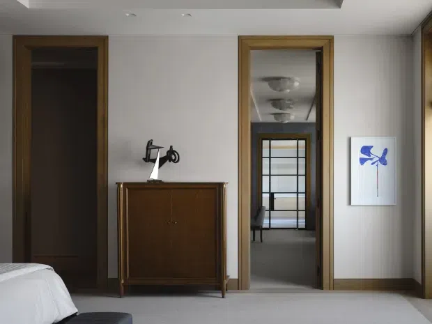 A refined bedroom interior with a neutral palette, grey carpet, white walls and ceiling, and dark timber door and trim details. A low vintage-style timber cabinet with a sculptural lamp sits against the wall, while an open doorway provides a view into an adjoining space with a glazed door and ceiling lighting. A small abstract artwork adds a blue accent.