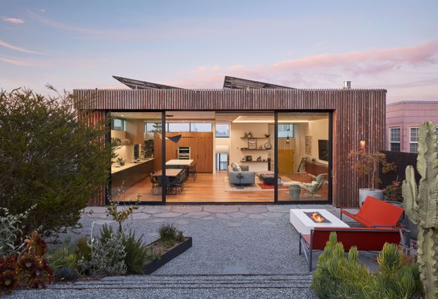 Contemporary single-storey residence viewed from a landscaped courtyard, with a strongly glazed living area opening to an outdoor terrace. The compact home has a rust-toned vertically ribbed exterior, angled roof elements, warm timber flooring and cabinetry, and a carefully designed desert-style garden with gravel paving, succulents, cacti, and outdoor seating around a fire table. The interior appears to include an open-plan living, dining, and kitchen area.