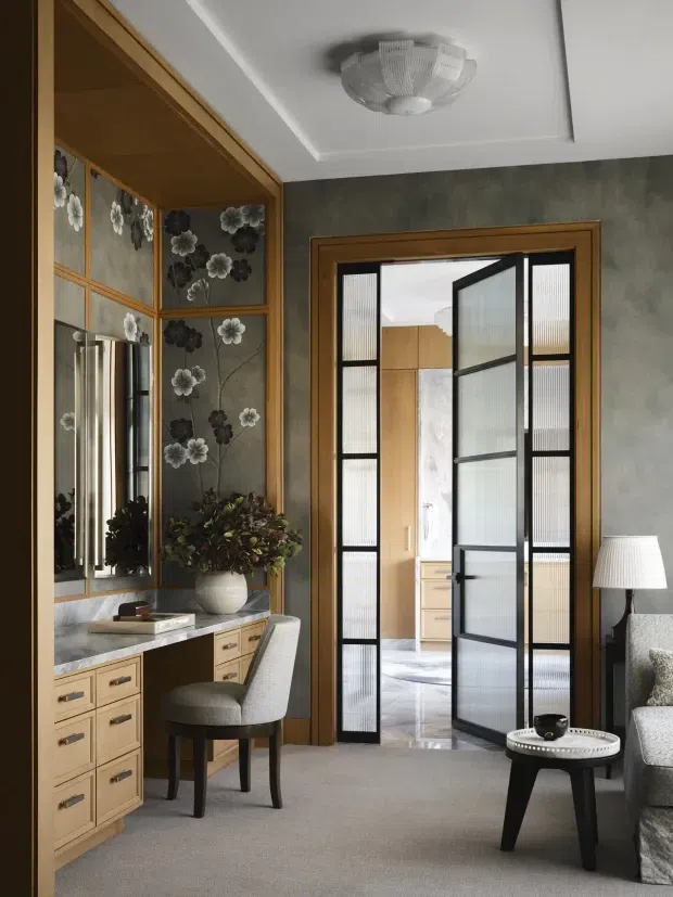 A refined bedroom interior featuring a built-in timber vanity and desk with drawers, a marble-look benchtop, mirrored cabinetry, and decorative floral glass panels. A grey upholstered chair sits at the vanity. Black-framed ribbed-glass double doors open directly to a bathroom or dressing area, while a partial sofa, side table, and lamp are visible on the right. The room has grey textured walls, neutral carpet, timber detailing, and a decorative flush-mount ceiling light.