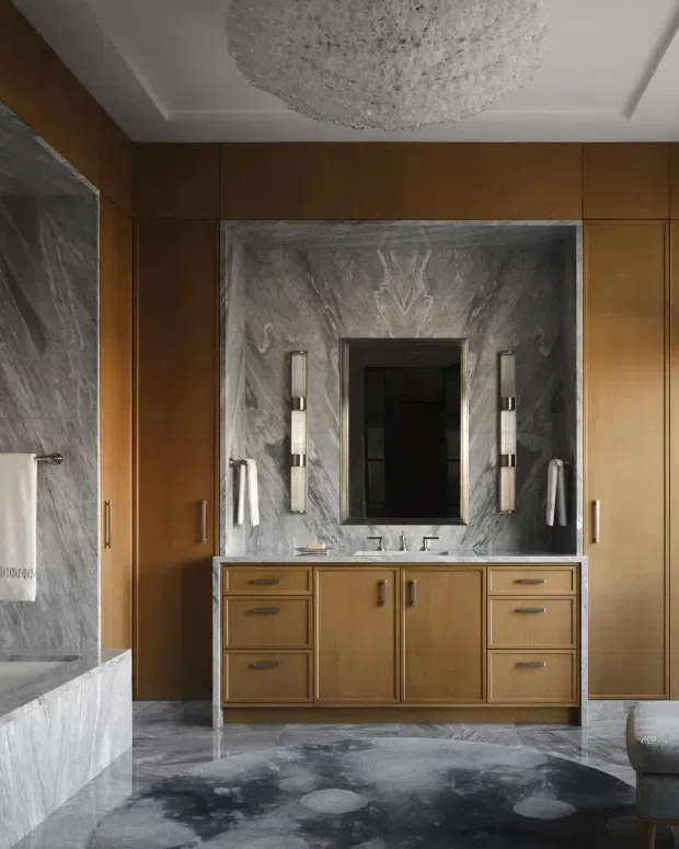 Luxurious bathroom interior featuring a large double vanity with warm wood cabinetry, grey veined stone wall and floor finishes, twin vertical wall sconces, integrated mirrors, and extensive floor-to-ceiling wood storage cabinetry. A freestanding or built-in bathtub is partially visible at left, while a sculptural textured chandelier and patterned rug add decorative detail.
