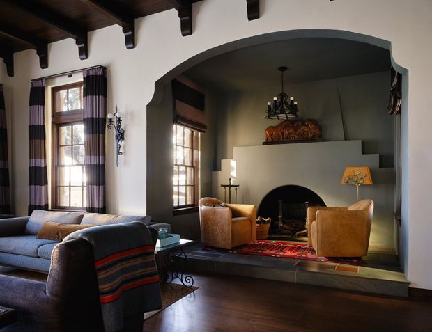 A richly detailed traditional living room centred on a large arched fireplace alcove. The room features dark exposed timber ceiling beams, deep wood flooring, tall multi-pane windows with striped drapery, a substantial blue-grey sofa, leather armchairs, and a raised hearth with a red patterned rug. The fireplace wall has stepped built-in detailing and a dark arched firebox, creating a warm, historic and lodge-like atmosphere.