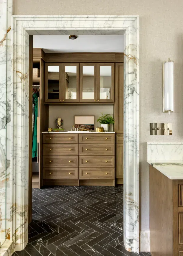 A refined walk-in wardrobe viewed through a dramatic marble-trimmed doorway. The space features dark timber cabinetry with mirrored upper doors, numerous drawers, open hanging storage, a decorative display shelf, and dark marble or stone herringbone flooring. A portion of an adjacent bathroom or vanity area with matching marble and timber joinery is visible at right.