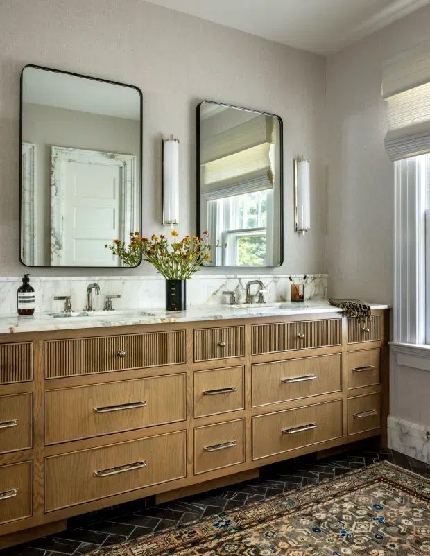 A refined bathroom vanity with a long fluted timber double vanity, white veined stone benchtop, two undermount sinks, paired black-framed mirrors, and wall-mounted vertical sconces. The room has textured neutral wallpaper, marble-look wall detailing, dark geometric floor tile, and a patterned runner rug. A window with layered blinds brings in natural light.