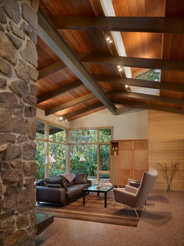A warm, rustic-modern living room with a dramatic timber-lined vaulted ceiling, exposed dark structural beams, and a substantial natural stone chimney or feature wall. Floor-to-ceiling timber-framed glazing overlooks dense greenery. The room contains a dark leather sofa, upholstered lounge chairs, a timber coffee table, built-in timber cabinetry, and layered area rugs over carpet.