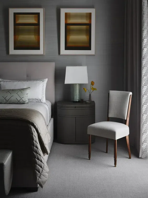 A refined, moody bedroom interior featuring a partially visible upholstered bed with layered white and patterned pillows and a charcoal quilt, a dark rounded bedside table, framed abstract artwork, a textured grey wall, and a light upholstered timber-legged chair beside floor-to-ceiling curtains and a screened window. The palette is predominantly grey, charcoal, white, and muted metallic tones, with small yellow floral accents.