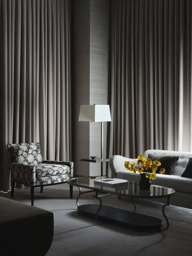A dark, refined living room with full-height pleated grey curtains, a textured grey sofa, patterned accent armchair, glass and dark metal coffee tables, and a sculptural floor lamp. The layered lighting and deep neutral palette create a sophisticated, moody atmosphere.